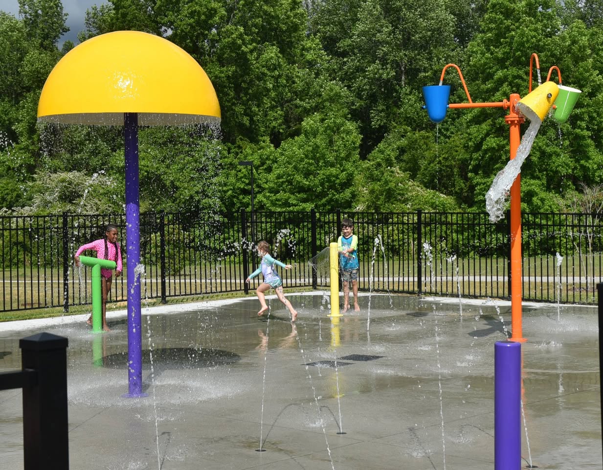 Children playing in a splash pad with colorful water features and fountains on a sunny day, surrounded by trees and a black fence.