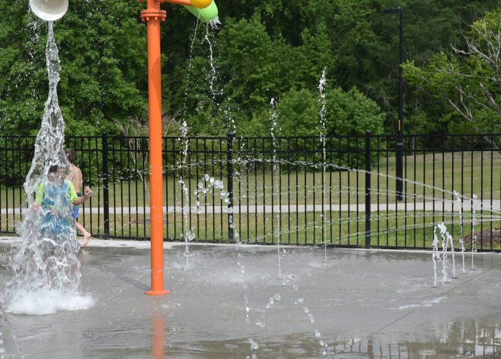 Children playing at a splash pad with water jets and an orange water feature, surrounded by a black metal fence and green trees in the background.