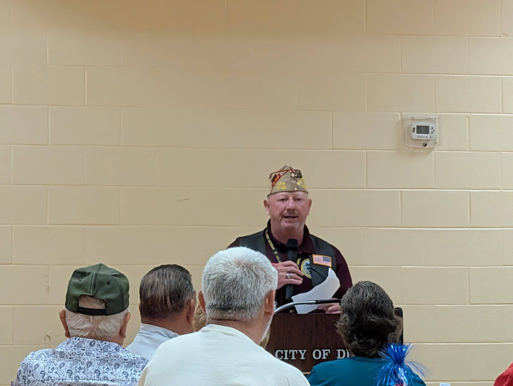 A man wearing a hat speaks at a podium to an audience in a room with beige walls.