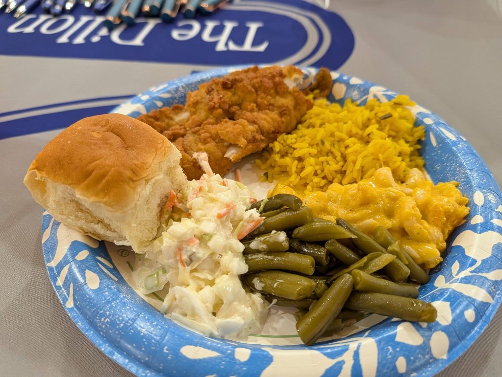 A paper plate with fried fish, yellow rice, macaroni and cheese, green beans, potato salad, and a dinner roll.