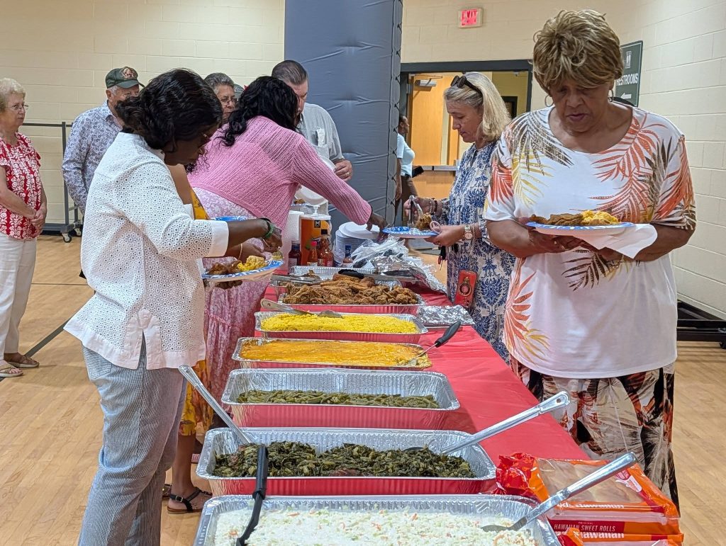 People serving themselves food from a buffet table covered with various dishes at a social gathering indoors.