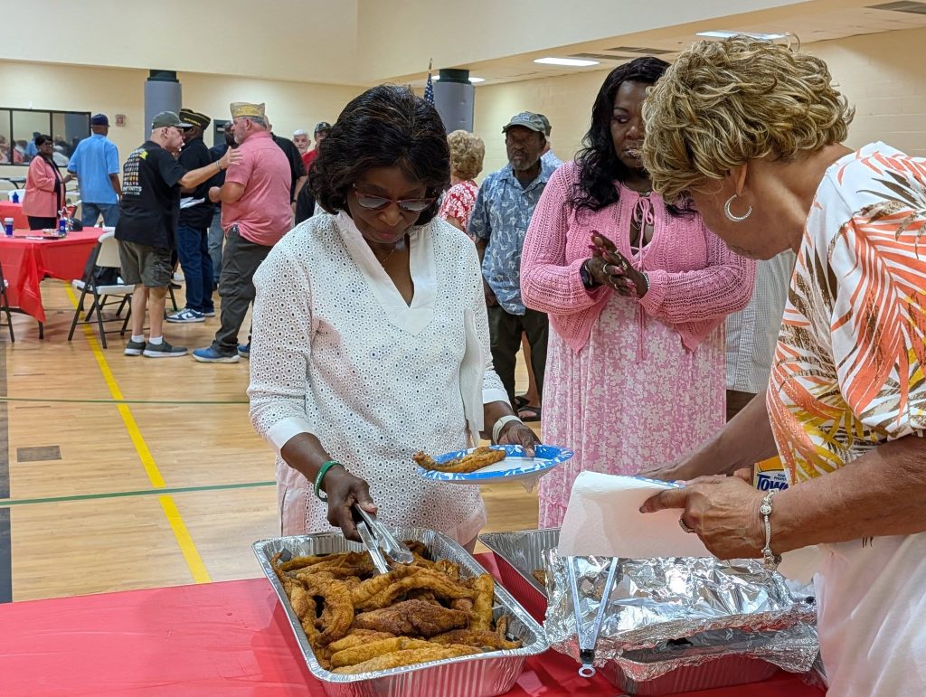 Women serving fried chicken from a tray at a community event, with people waiting in line inside a gymnasium.