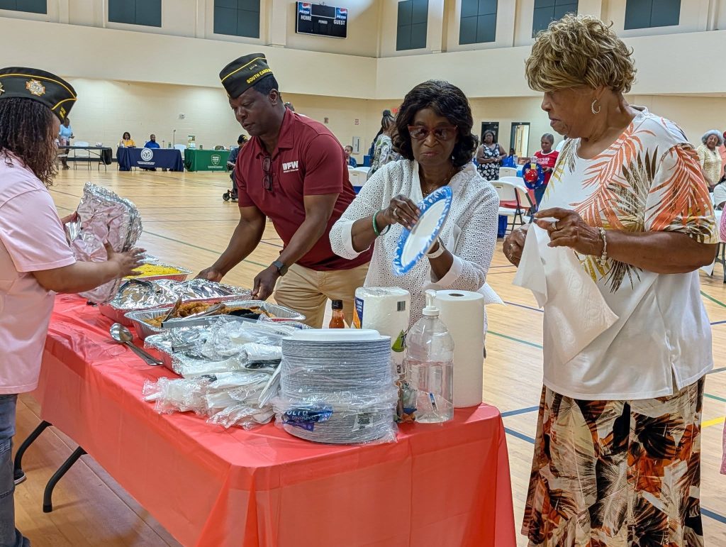 Four people standing at a red-covered table serving and preparing food in a large indoor community center, with more tables and people in the background.