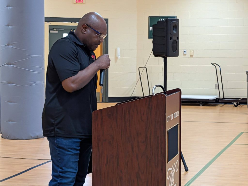 A man stands at a podium speaking into a microphone in a gymnasium, with a speaker and some equipment visible in the background.