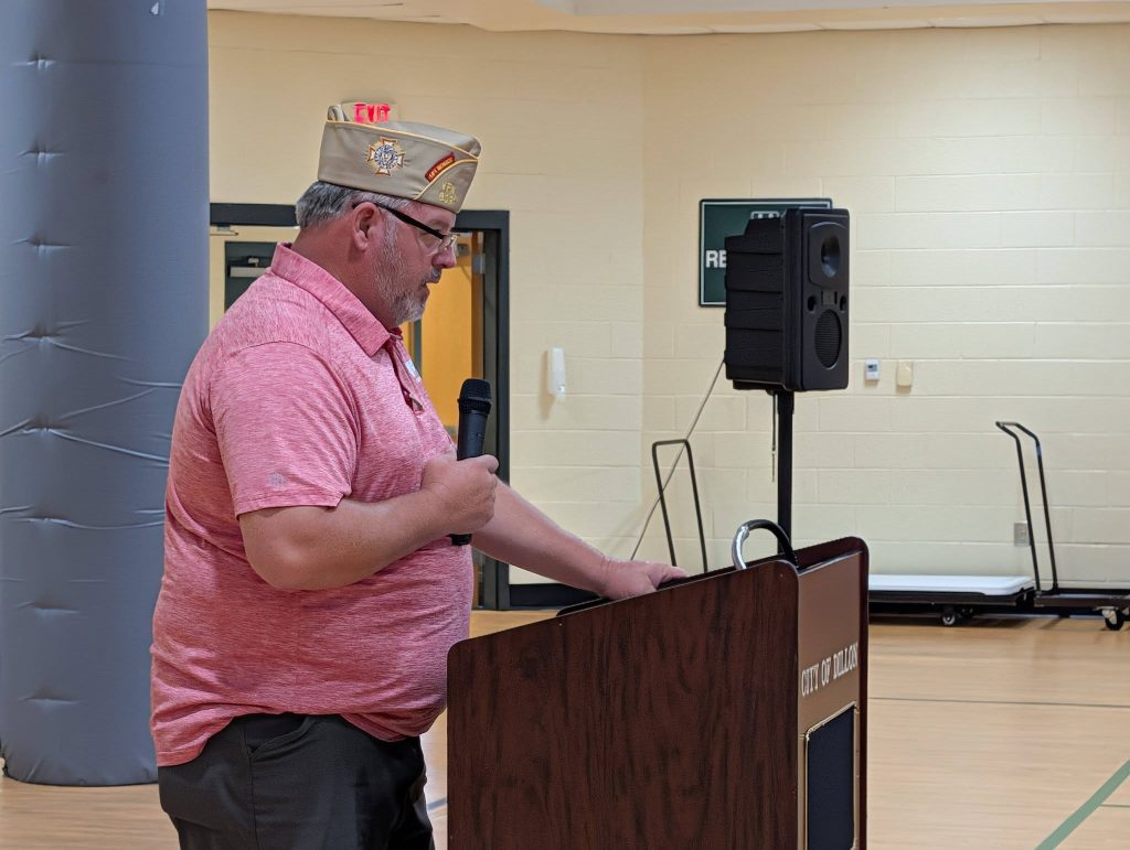 A man wearing a pink shirt and hat speaks at a podium in a large indoor room with a speaker in the background.