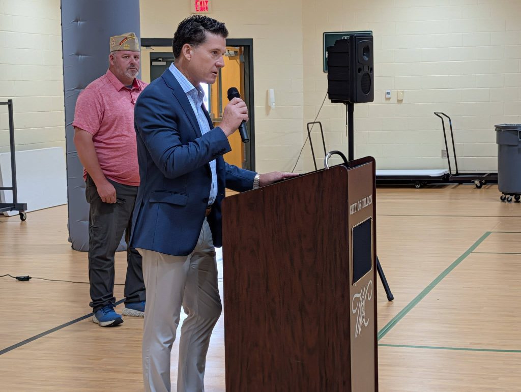 Man in suit speaking at podium with microphone in a gymnasium, another man in casual clothes stands in the background near a speaker.