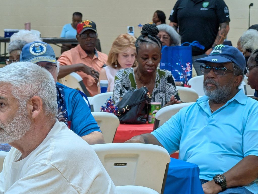 People sit at tables in a community gathering, talking and socializing, with decorations and drinks visible on the tables.