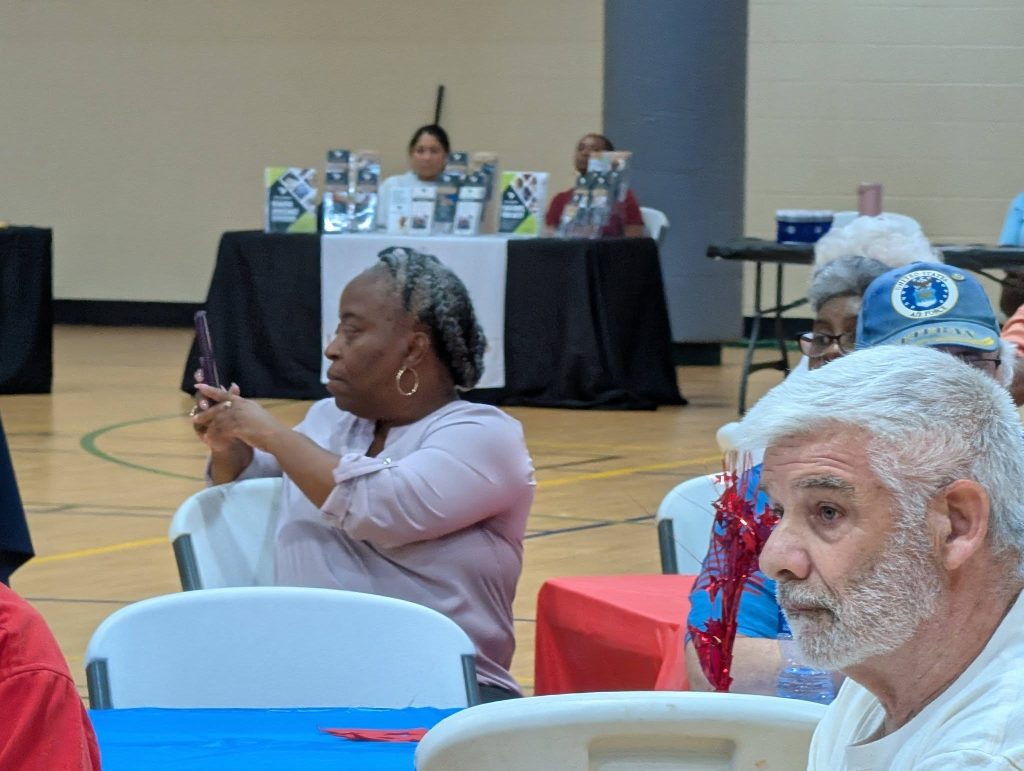 Several older adults sit at tables in a community center. One woman takes a photo with her phone, while others listen. A panel sits at a table in the background.