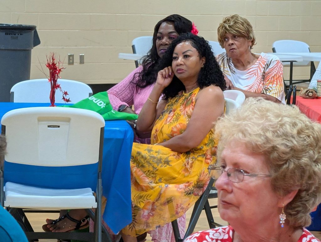 Four women sit at decorated tables in a brightly lit room, appearing to listen attentively during an indoor event or gathering.