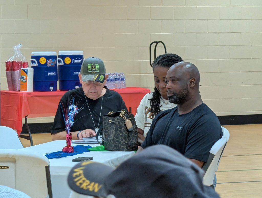 Three people sit at a table in a community center, one wearing a military cap, with coolers and a red tablecloth in the background.