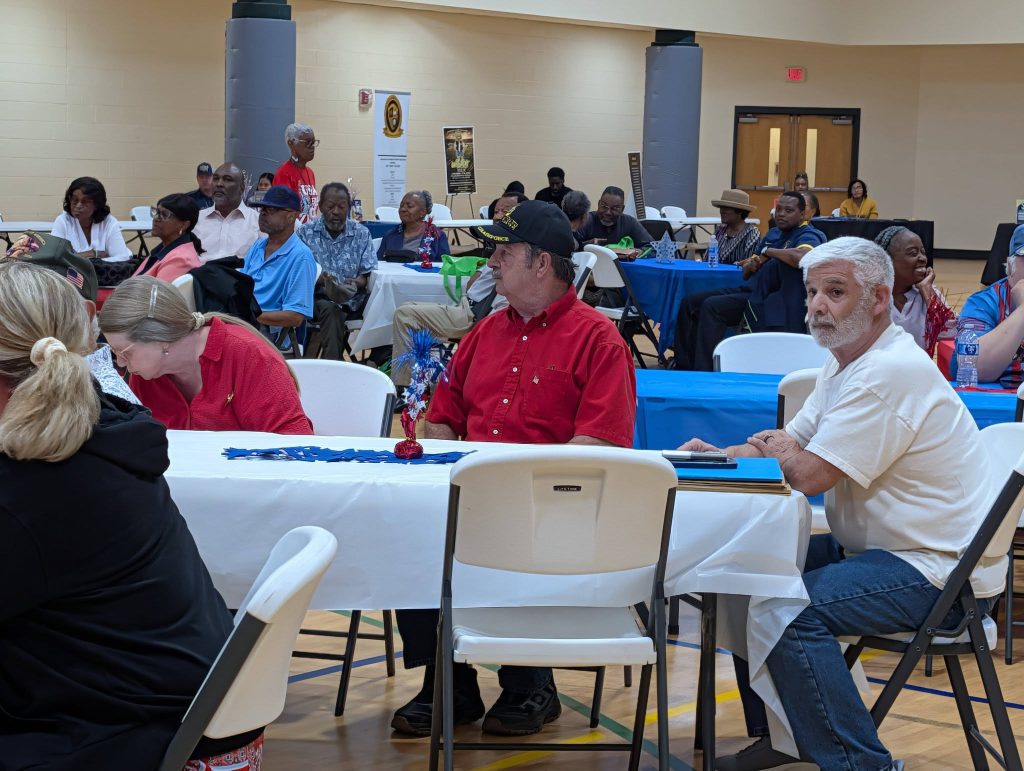 People seated at tables in a community hall, engaged in conversation and activities. The room is filled with round and rectangular tables, covered with white and blue tablecloths.