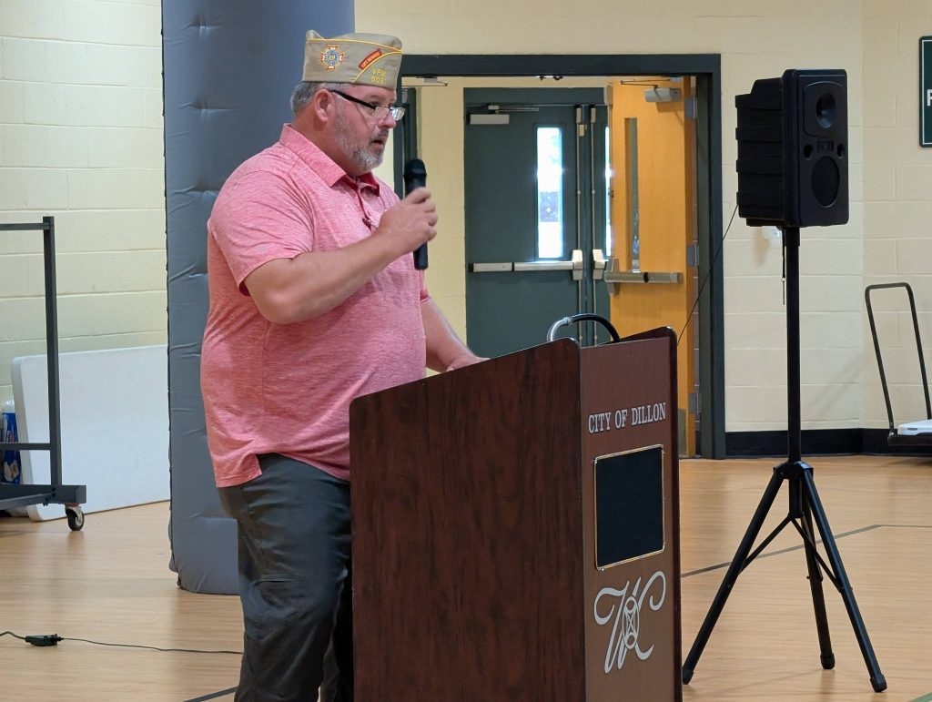 Man wearing a beige hat and pink shirt speaks into a microphone at a wooden podium inside a building, with doors and a speaker visible in the background.