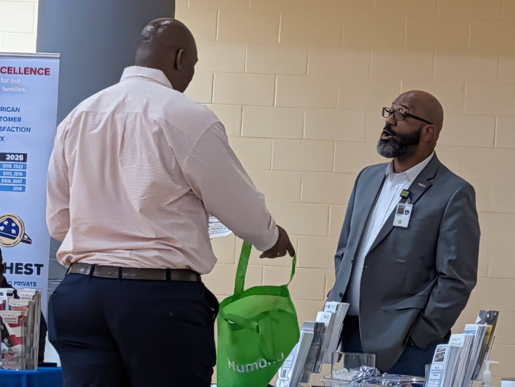 Two men having a conversation indoors; one is holding a green tote bag, while the other stands behind a table wearing a suit and name badge.