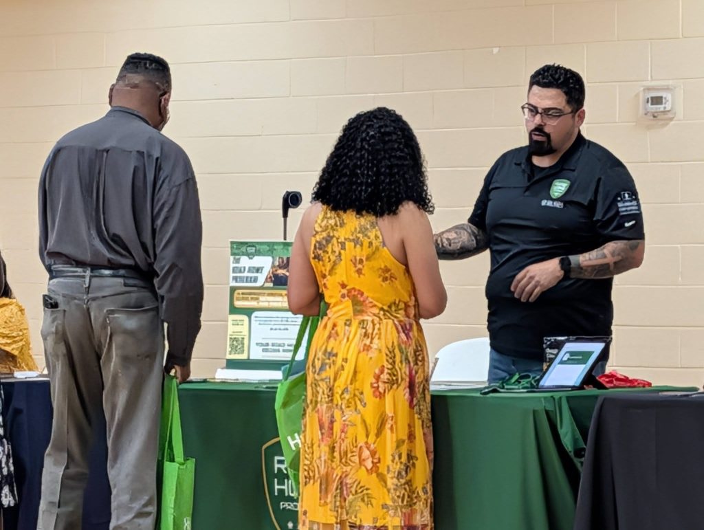 Three people stand at an informational booth with a green tablecloth, talking. One woman in a yellow dress faces the table, two men are on either side.