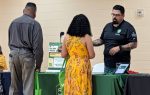 Three people stand at an informational booth with a green tablecloth, talking. One woman in a yellow dress faces the table, two men are on either side.