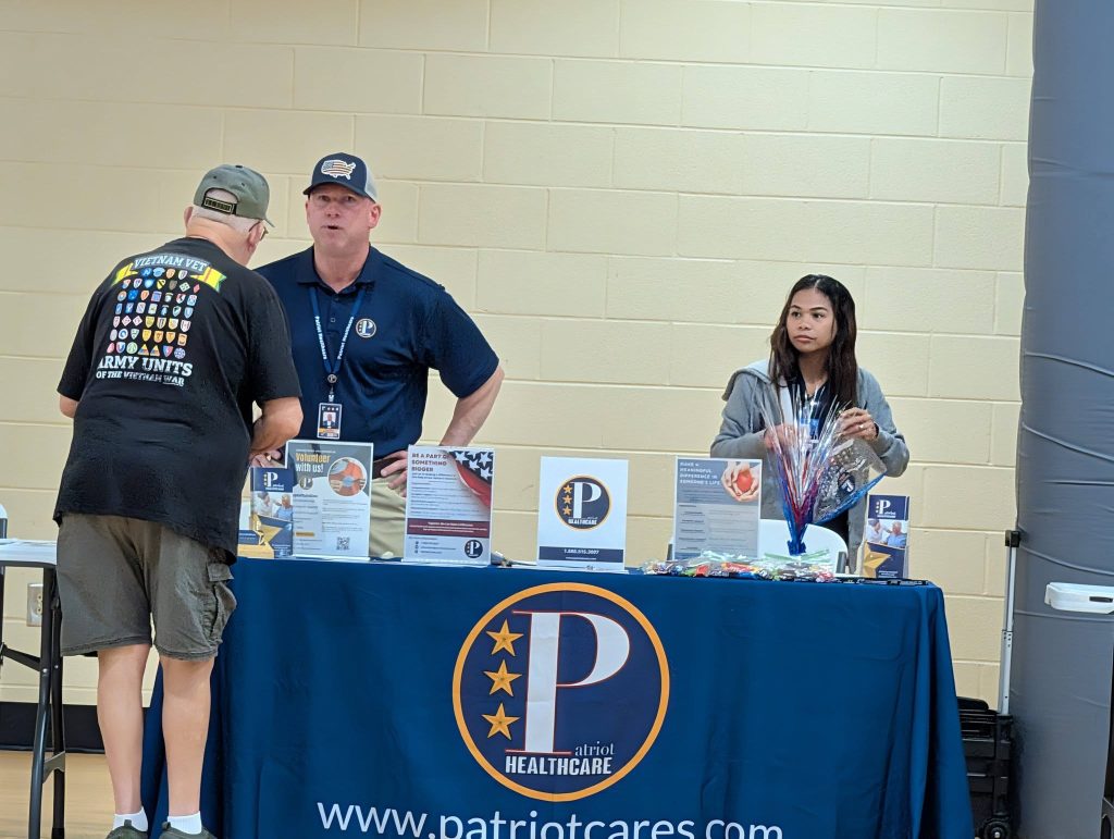 Three people stand at an informational booth with a blue tablecloth displaying "PatriotCares" logo and pamphlets, in front of a beige wall.