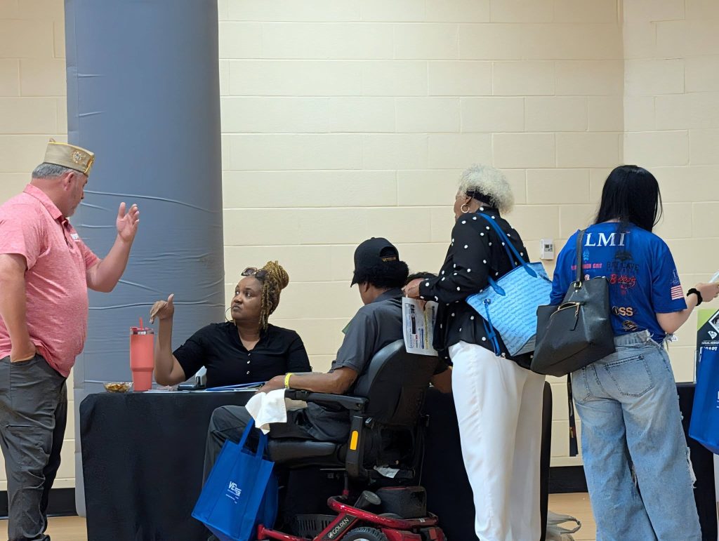 People gather at a table for assistance; one person sits in a mobility scooter, while others stand and converse with individuals seated behind the table.