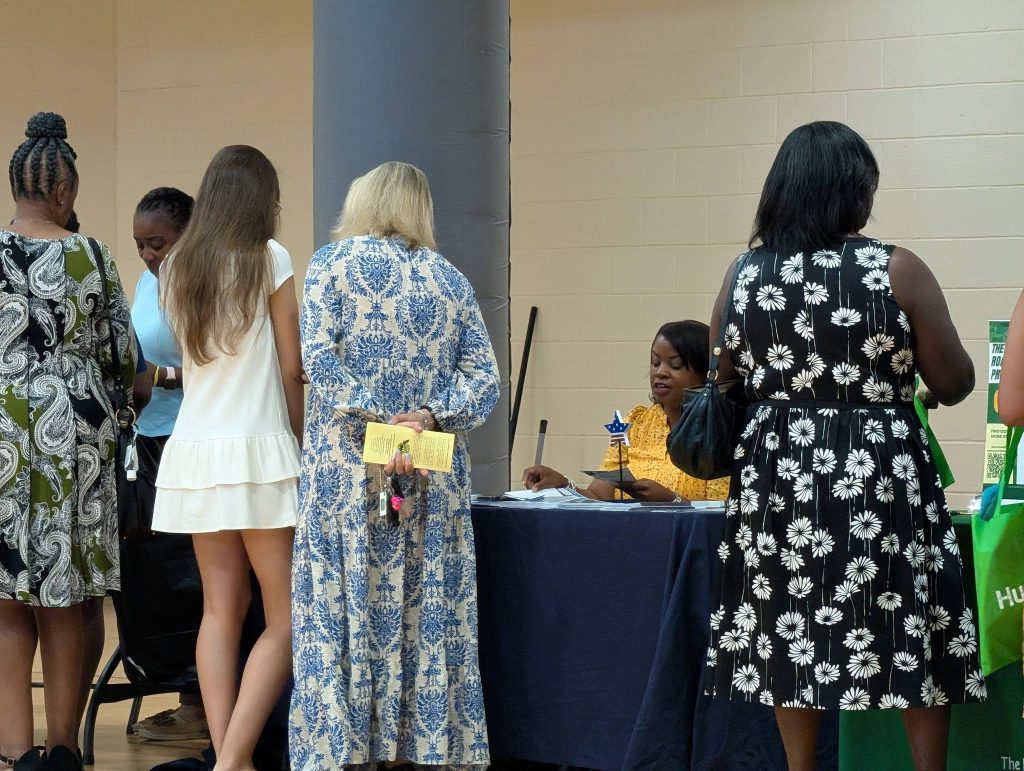 Several women stand in line at a table where another woman is seated, possibly for registration or information at an indoor event.