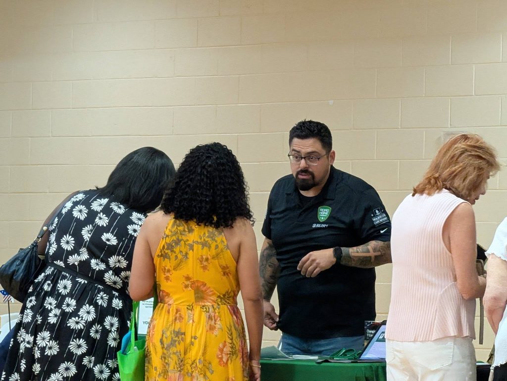 A man at a table talks to three women standing in front of him at an indoor event.