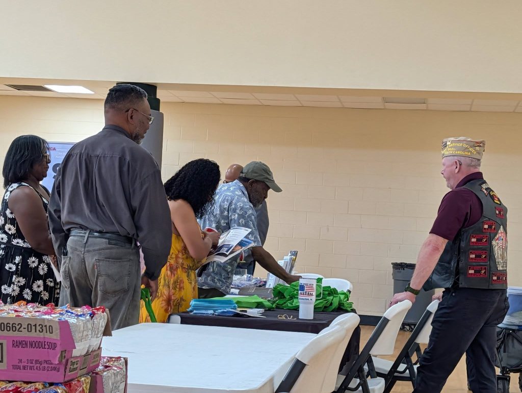 Several people stand around a table indoors, looking at items on display. The setting appears to be a community event or gathering.