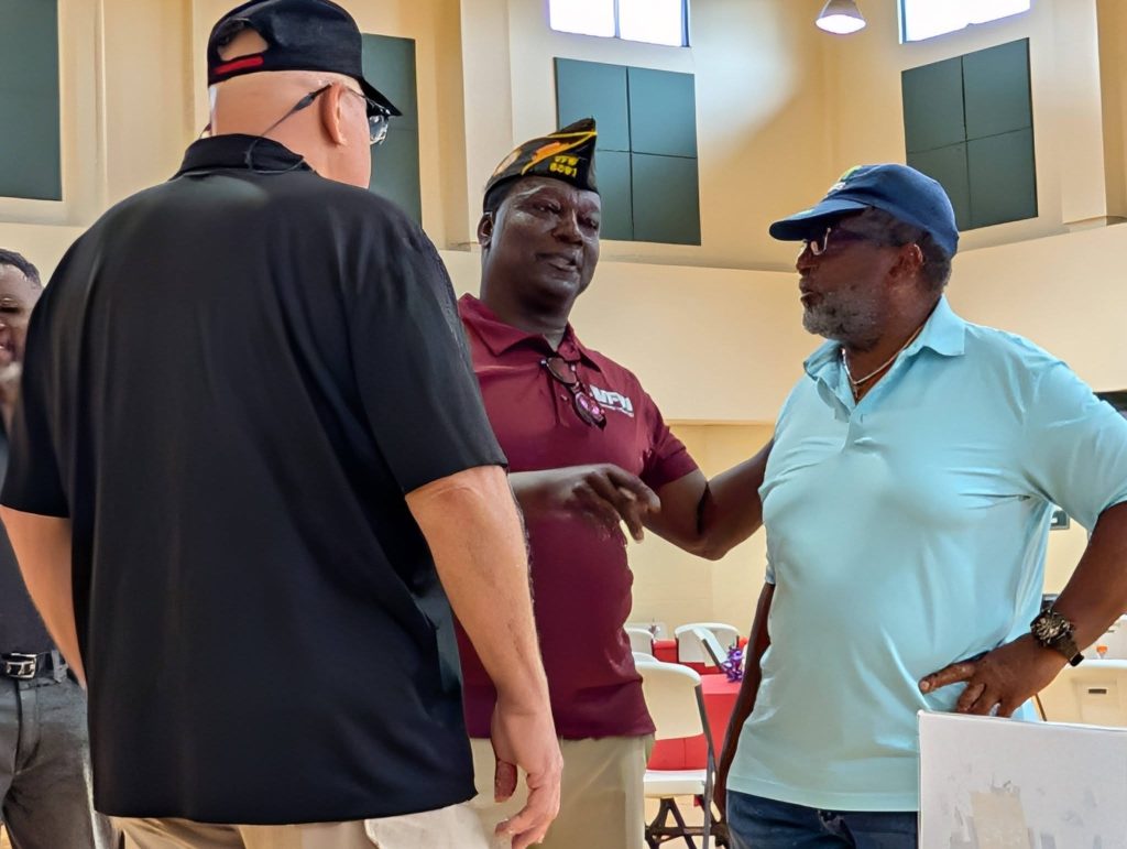 Three men wearing hats stand indoors, conversing. One man wears a military cap and maroon shirt, while the others wear casual shirts and baseball caps.
