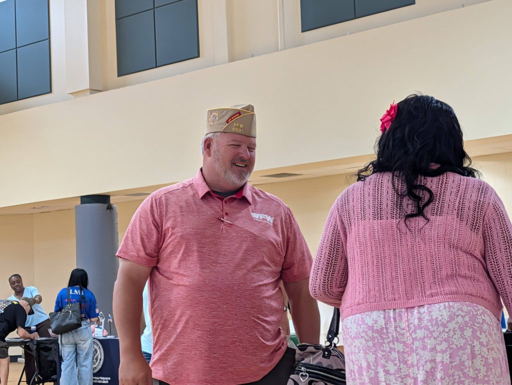 A man in a tan hat and pink shirt talks to a woman in a pink outfit indoors at an event.