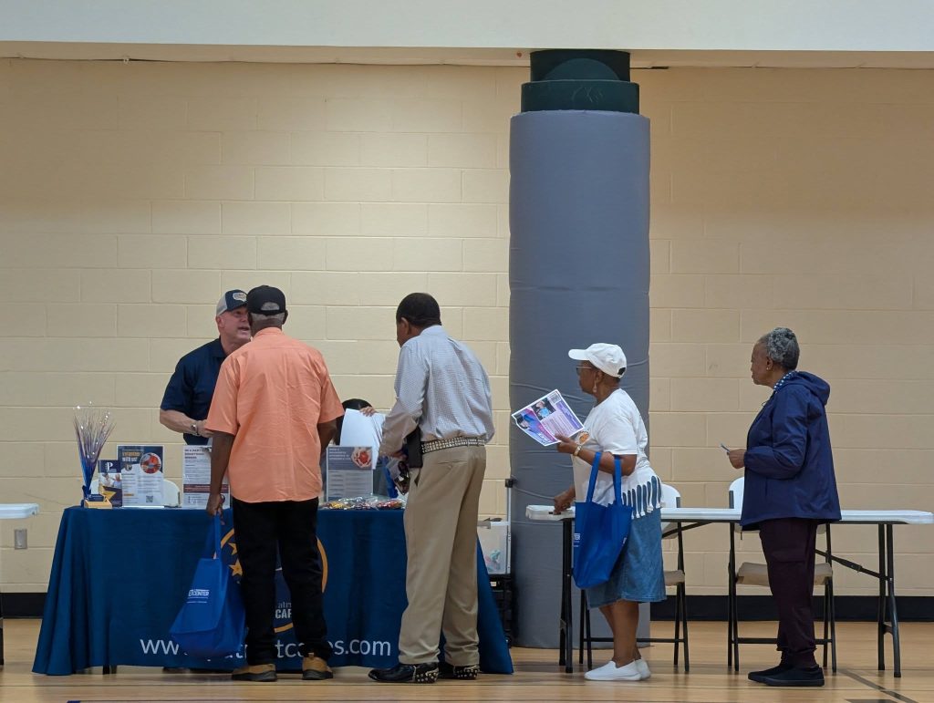 Several people stand at an indoor event table, engaging with vendors and collecting informational materials in a community center setting.
