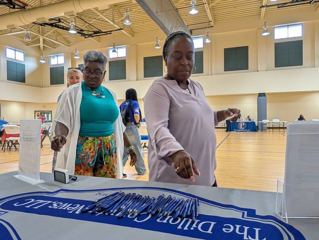 Two women stand at a table with pens and a blue banner inside a gymnasium, appearing to select items during an indoor community event.