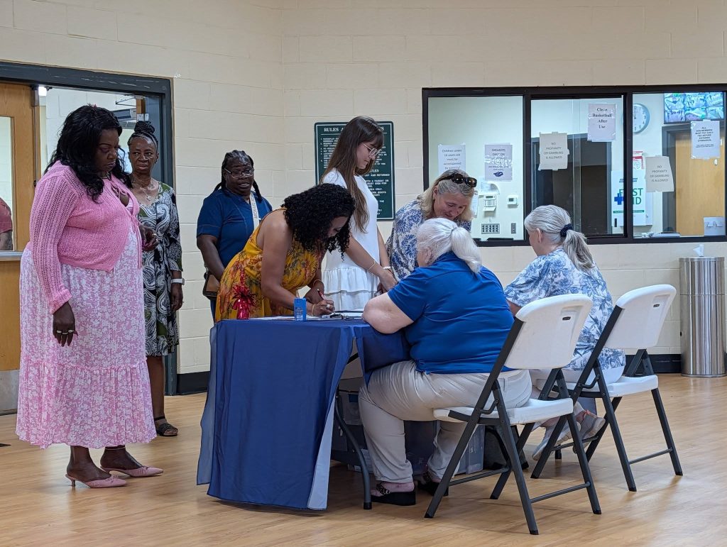 Several people stand and sit around a table indoors, signing papers and interacting, possibly at an event registration or check-in area.