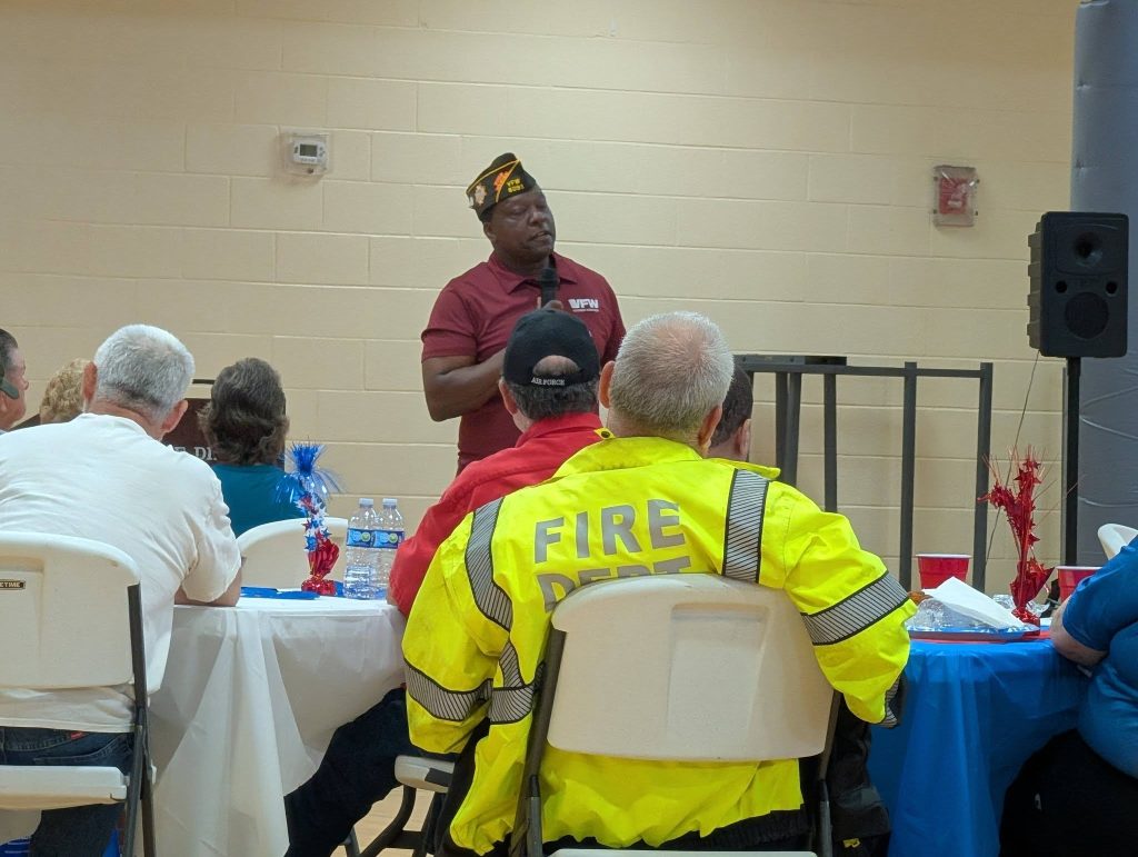 A man in a uniform speaks to seated people, including one wearing a bright yellow "FIRE" jacket, in a community room setting.