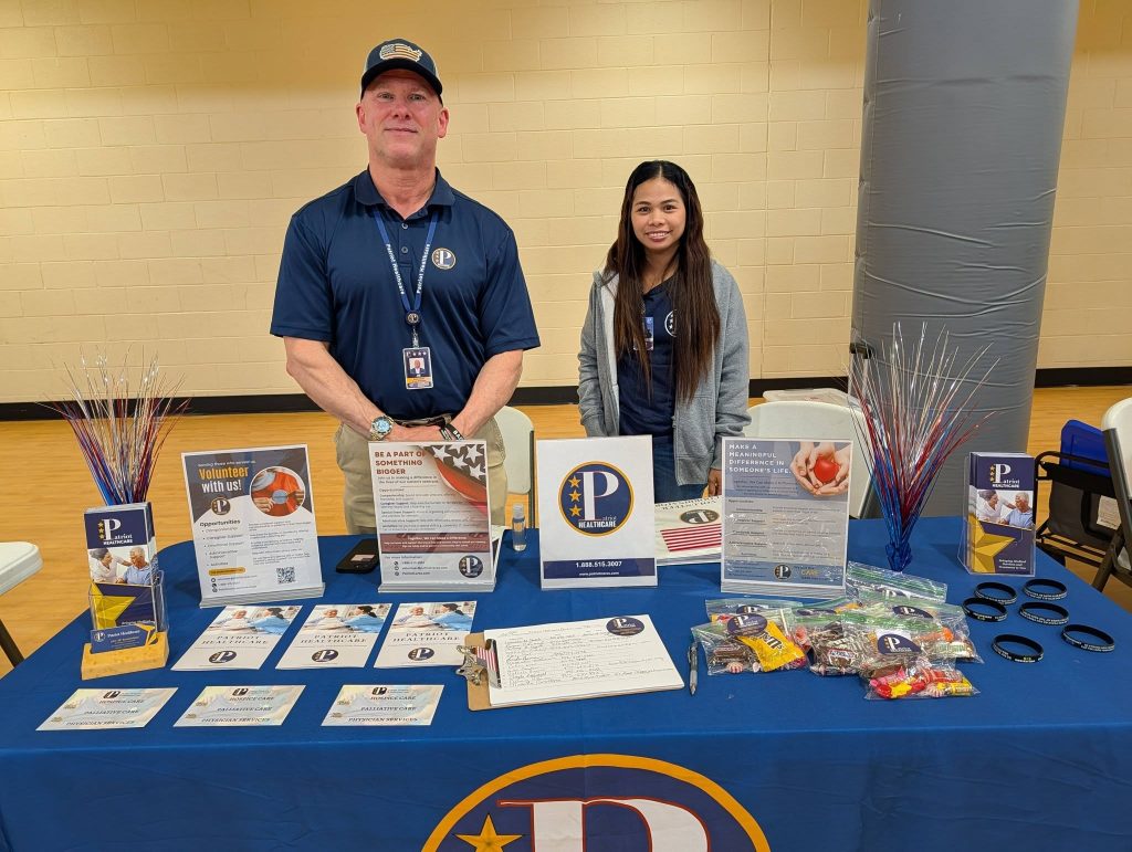 Two people standing behind an informational table with pamphlets, flyers, and small giveaway items, promoting a community organization indoors.