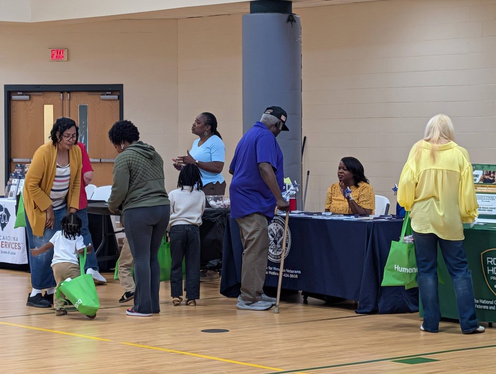 People standing and talking at indoor tables during a community event; some are holding green bags and others are seated behind the tables.