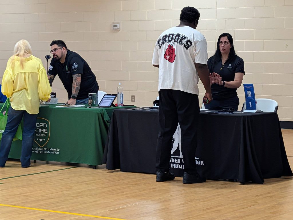 People interacting at an indoor event with tables, laptops, and informational displays. One person wears a white "Crooks" shirt while speaking to a woman behind a table.