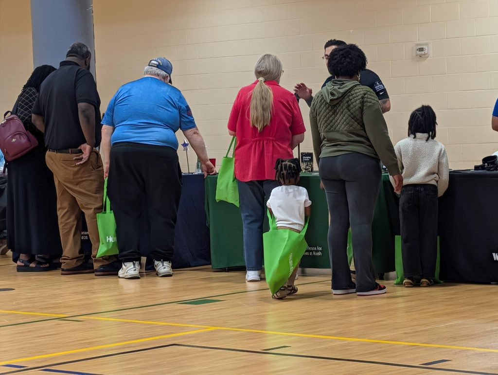People of various ages stand in line indoors, holding green tote bags, in front of a table at what appears to be a community or school event.