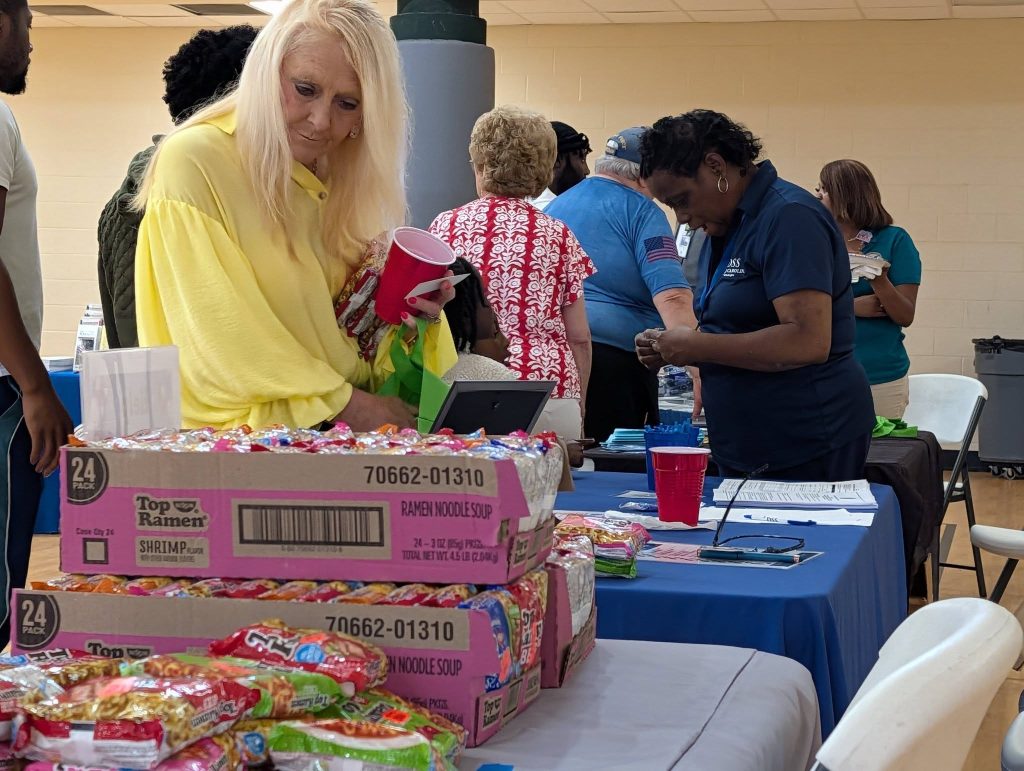 A woman in yellow looks at ramen noodle packs on a table, with several people gathered around booths in a busy indoor setting.