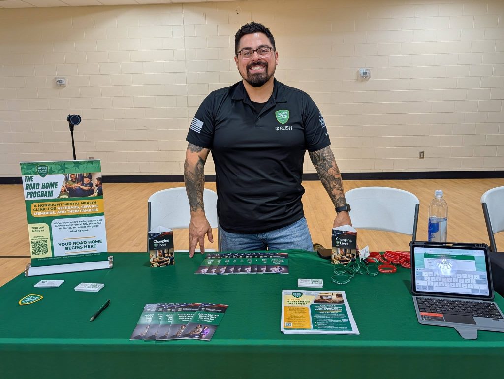 Smiling man with tattoos stands behind a green-covered table displaying brochures, flyers, and a laptop at an indoor event or expo.
