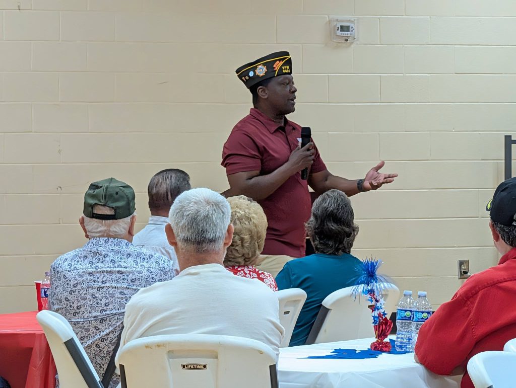 A man wearing a military veterans cap speaks into a microphone while standing in front of a seated audience in a community hall.