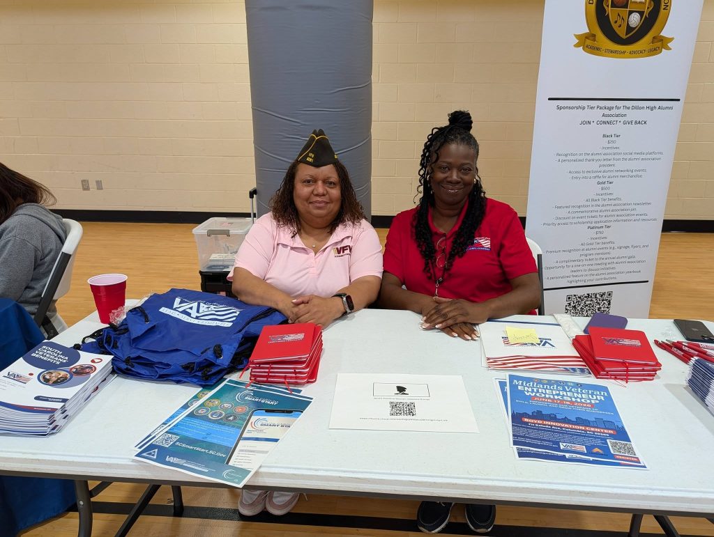 Two women sit at a table with informational brochures, tote bags, and pamphlets, participating in an indoor community or informational event.