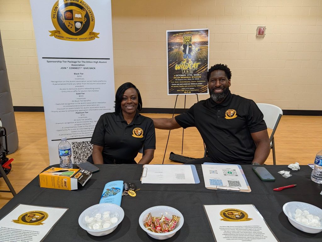 Two people sitting at an information booth with brochures, snacks, and promotional materials, both wearing matching black shirts with a yellow logo, smiling at the camera.