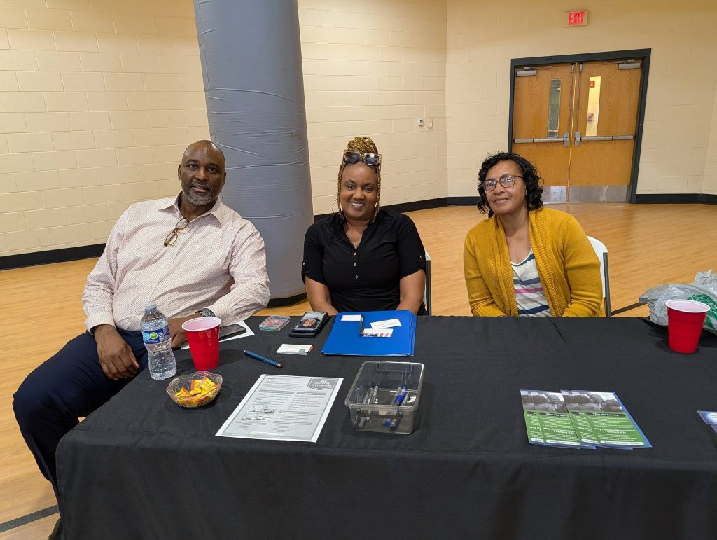 Three people sit behind a table with snacks, papers, and informational materials, smiling in a brightly lit indoor setting.