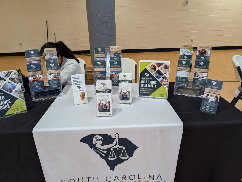 A display table with brochures and flyers about South Carolina legal services, featuring a logo with scales of justice and the state outline.