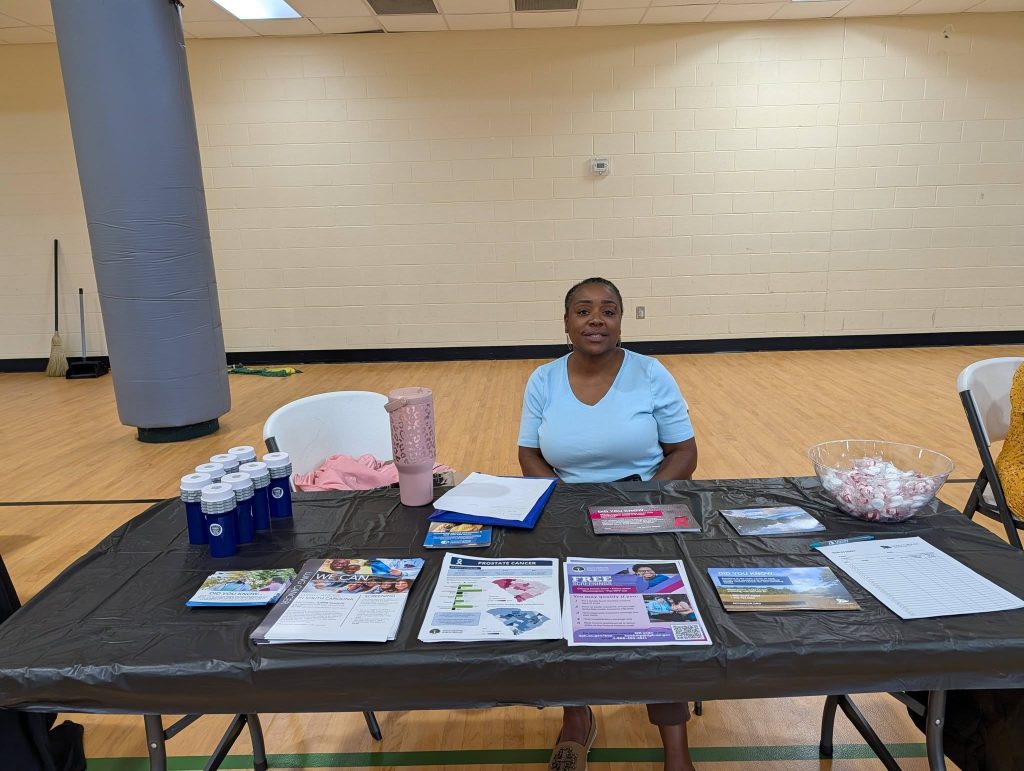 A woman sits behind a table with informational pamphlets, water bottles, and small items, in a gymnasium setting.