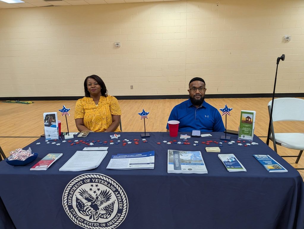 Two people sit behind a table covered with brochures and promotional materials, in a gymnasium or event hall. A seal or emblem decorates the tablecloth.