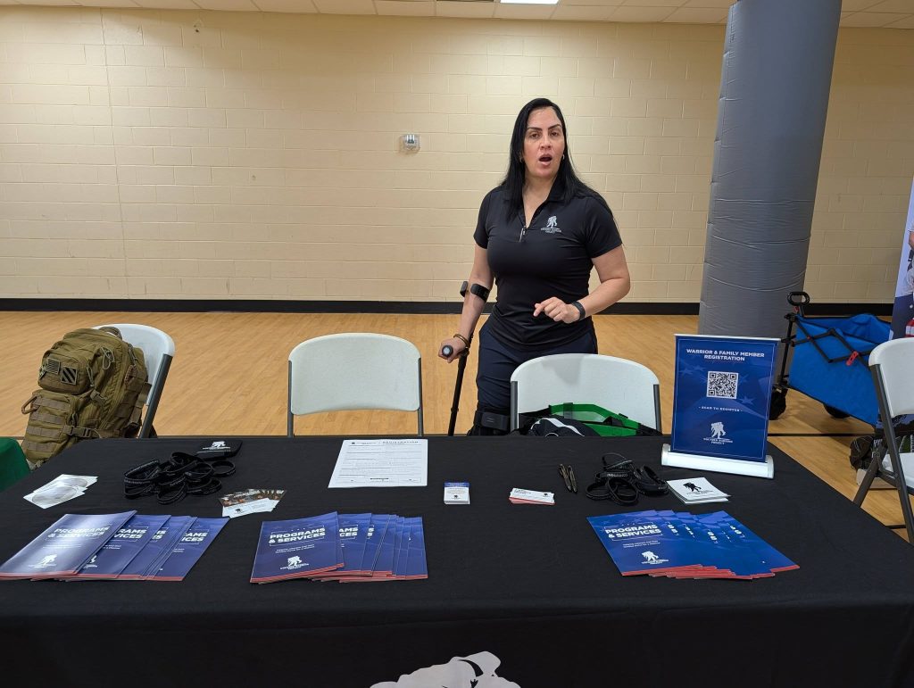 A woman stands behind a table with brochures and promotional items at an indoor event, ready to assist visitors.