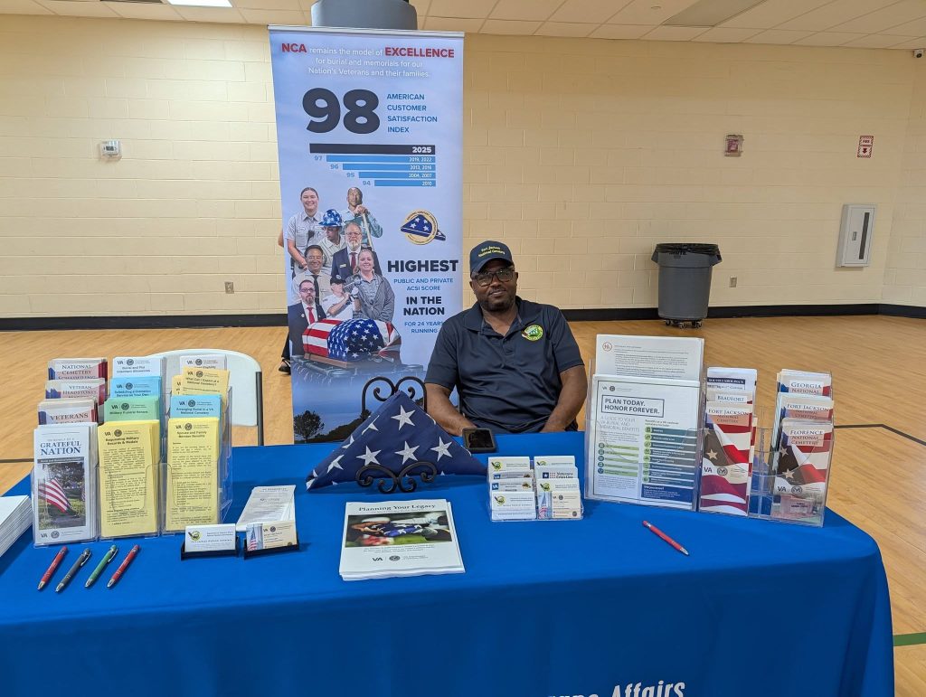 A man sits behind an informational table with brochures, pamphlets, and flags at a veterans affairs event indoors.