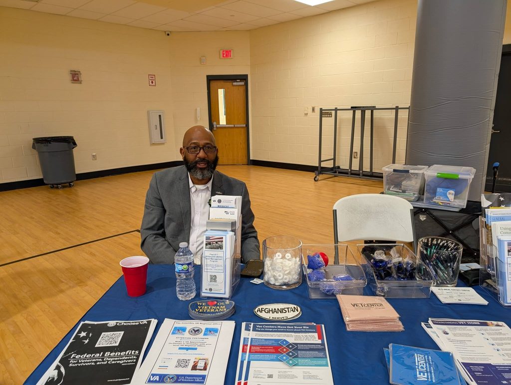 A man sits at a table with informational brochures, water, and promotional items in a gymnasium or event space.