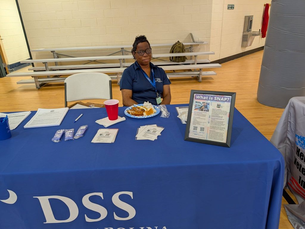 A woman sits behind a DSS information table with brochures, snacks, and a framed sign in a gymnasium setting.