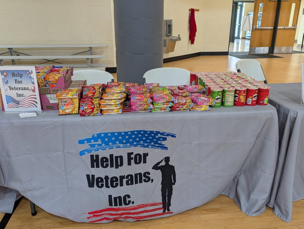 A table covered with snacks and a cloth that reads "Help For Veterans, Inc." is set up indoors, likely for a community or charity event.