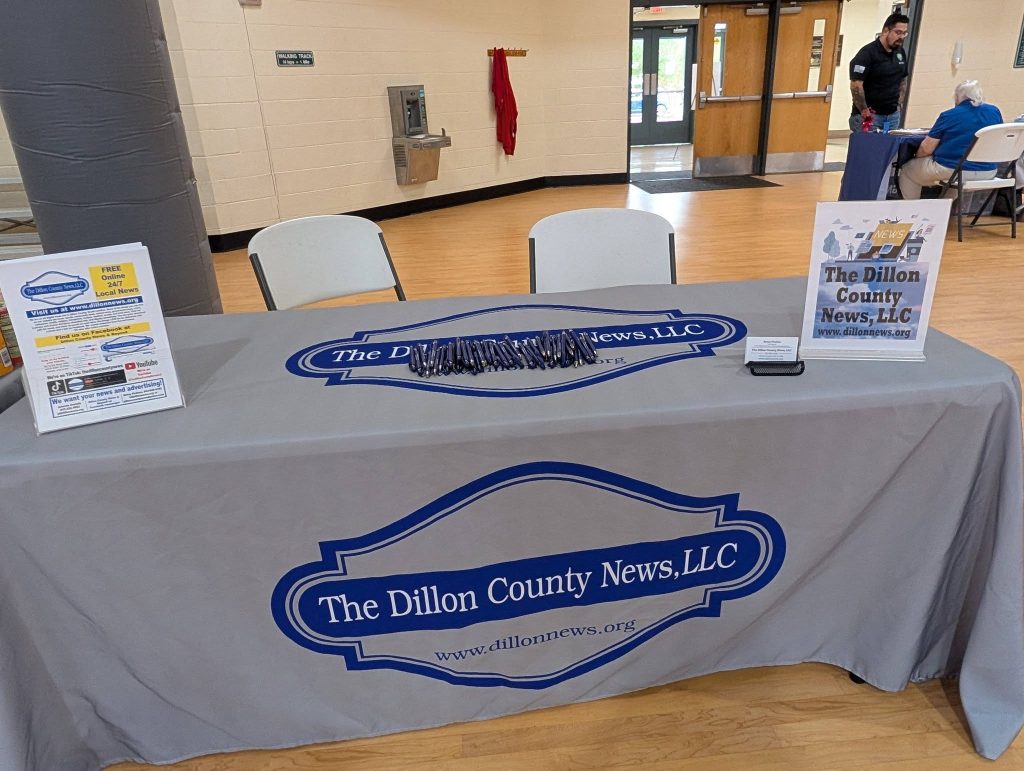 A table with a gray cloth labeled "The Dillon County News LLC," brochures, and chairs set up in an indoor venue.