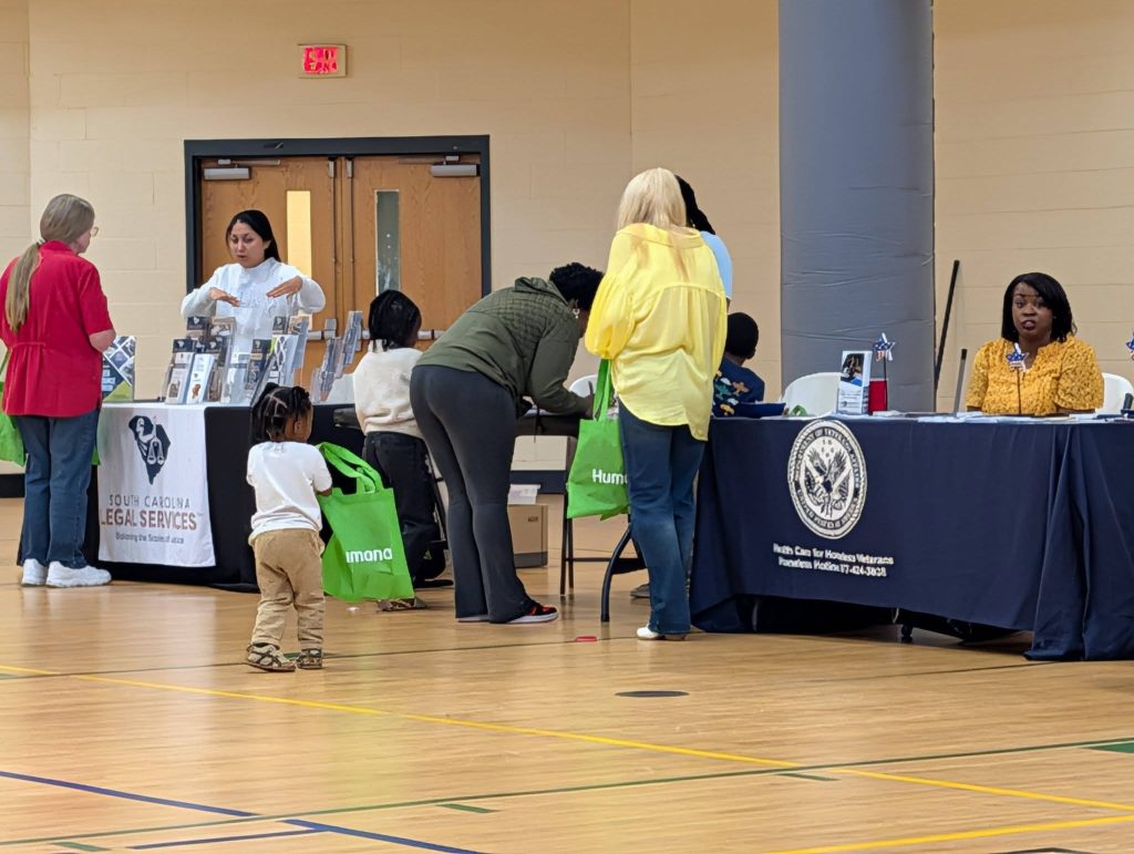 People visit tables with informational displays at an indoor event; a child holding a green bag walks toward adults who are talking with representatives.